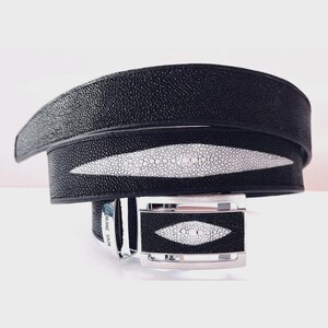 May include: Black belt with a white stingray skin inlay. The belt has a silver buckle with a matching stingray skin design. The words "GENUINE SKIN" are visible on the belt.