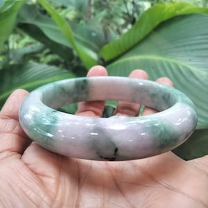 May include: A smooth, polished bangle bracelet crafted from natural stone, featuring a rounded profile with a marbled appearance. The surface displays a blend of translucent pale lavender and deep forest green patches throughout the circular band.