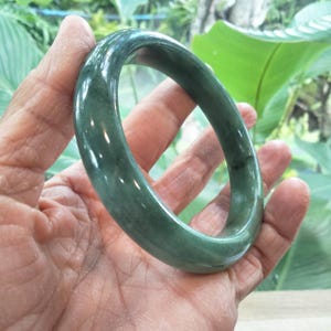 May include: A smooth, polished jade bangle bracelet featuring a deep forest green colour with subtle, darker mottled inclusions throughout the stone. The circular jewellery piece has a rounded profile and a glossy, reflective surface finish.