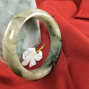 May include: A jade bangle bracelet with a mix of green and white tones. The smooth, polished surface of the bracelet is highlighted against a red background. A white flower with a yellow stem is nestled inside the bracelet.