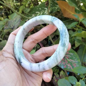 Puede incluir: Una pulsera de jade con aspecto veteado. La pulsera presenta una mezcla de tonos blancos, verde claro y verde más oscuro. La superficie lisa y pulida refleja la luz, resaltando las variaciones naturales de la piedra.