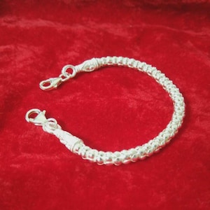 Può includere: Un braccialetto d'argento con un design strutturato e perlato. Il braccialetto presenta una serie di piccole perle rotonde collegate tra loro, con una chiusura a moschettone. Il braccialetto è esposto su una superficie di velluto rosso.