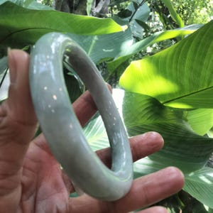 以下が含まれることがあります： 淡い半透明の緑色の翡翠で作られた、滑らかで磨き上げられたバングルブレスレット。円形のバンドは丸みを帯びた形状で、光を反射する光沢のある仕上げが施されており、石の自然な模様と柔らかな色合いが際立っています。
