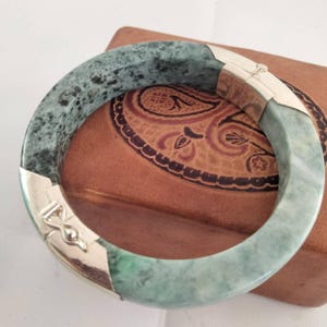 Könnte beinhalten: Ein Jade-Armreif mit silberfarbenen Metallakzenten. Das Armband hat einen gesprenkelten grün-weißen Stein und einen sicheren Verschluss. Der Armreif liegt auf einer braunen Lederoberfläche.
