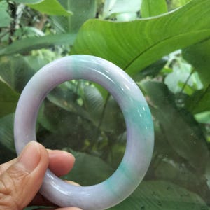 Puede incluir: Un brazalete circular y liso hecho de piedra pulida. La pieza presenta una apariencia translúcida con una mezcla de variaciones de color lavanda suave y verde vibrante distribuidas por toda la estructura redondeada y sólida.