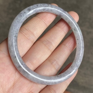 以下が含まれることがあります： 丸い、明るい灰色の翡翠のバングルブレスレット。 ブレスレットは、色と質感に微妙な変化がある、滑らかで研磨された表面を持っています。 翡翠は、より濃い灰色の斑点のある淡い灰色です。