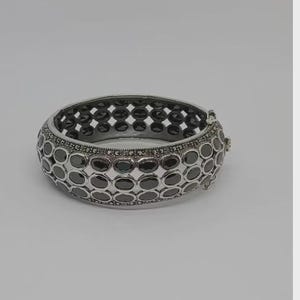 Può includere: Bracciale rigido a cerniera argentato con un motivo ripetuto di pietre circolari grigio scuro. Il bracciale ha una chiusura sicura e un interno traforato. Un elegante gioiello.