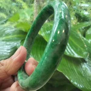 Puede incluir: Un brazalete rígido liso y pulido hecho de jade verde. El accesorio circular presenta un perfil redondeado con variaciones de color naturales, mostrando tonos esmeralda profundos mezclados con inclusiones nubosas blancas translúcidas.