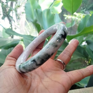 May include: A jade bangle bracelet with a smooth, polished surface. The bracelet features a mix of white, light green, and dark green colors. The jade is held in a hand, with a silver ring on the finger.