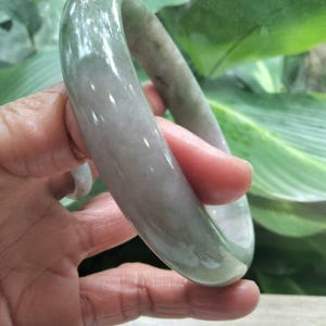 以下が含まれることがあります： 半透明の淡い緑色の翡翠で作られた、滑らかで磨き上げられたバングルブレスレット。円形のアクセサリーは丸みを帯びた形状で、石の表面全体に明るい灰色から柔らかな緑色までの微妙な色の変化が見られます。