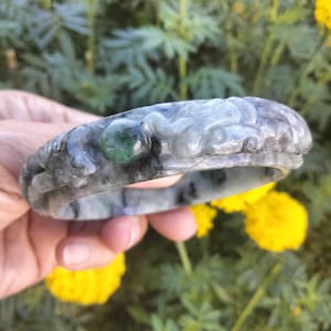 May include: A carved jade bangle bracelet in shades of gray and green. The bracelet features intricate dragon or mythical creature designs. A small, round, green jade stone is set into the bracelet. The background is blurred with yellow flowers and green foliage.