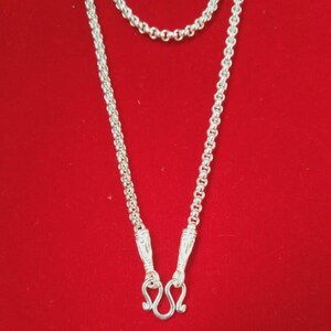 Könnte beinhalten: Eine silberne Halskette mit rundem Gliederkettendesign. Die Kette endet in zwei dekorativen, texturierten Metallkappen, die zu einem gebogenen Hakenverschluss führen. Die metallische Oberfläche weist durchgehend ein helles Finish auf.