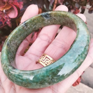 Può includere: Un braccialetto di giada verde lucido. Il braccialetto ha un colore verde screziato con macchie di verde più scuro.