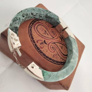 Peut inclure: Un bracelet jonc en jade avec un fermoir en argent. Le bracelet est de couleur vert marbré et repose sur une boîte en cuir marron avec un motif cachemire.