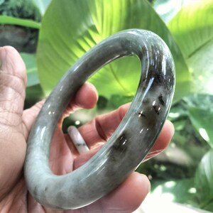 以下が含まれることがあります： 半透明の淡いグレーグリーンのベースカラーを持つ、磨かれた石で作られた滑らかで丸みを帯びたバングルブレスレット。表面には、濃いチャコールグレーのインクルージョンと斑点模様がはっきりと見られ、自然でマーブル状の外観を作り出しています。