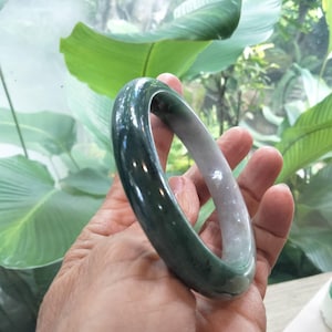 以下が含まれることがあります： 深いフォレストグリーンから半透明のペールグレーへと変化するグラデーションが特徴の、滑らかで磨き上げられたバングルブレスレット。円形のアクセサリーは丸みを帯びた輪郭を持ち、光を反射する光沢のある表面がその曲線に沿って輝いています。