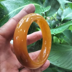May include: A smooth, polished bangle bracelet crafted from translucent golden-yellow stone. The circular accessory features a rounded profile with a glossy, reflective surface that catches the light, showcasing a consistent warm amber hue throughout.