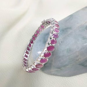 May include: A silver bangle bracelet with oval-cut, pink gemstones. The bracelet is set with multiple gemstones, and the clasp is visible. The jewelry is displayed on a white surface.