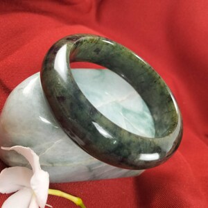 May include: A dark green jade bangle bracelet. The smooth, polished bracelet is resting on a light green stone against a red background. A white flower is in the foreground.