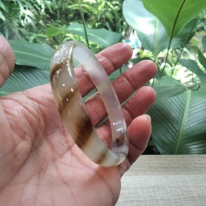 May include: A smooth, translucent bangle bracelet featuring a polished finish. The circular accessory displays a distinct pattern of swirling brown and cream-colored bands that wrap around the entire circumference of the solid, rounded band.