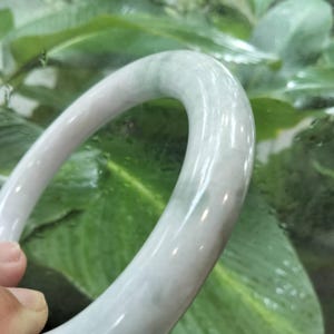 以下が含まれることがあります： 淡い半透明の石で作られた滑らかで磨き上げられたバングルブレスレット。丸みを帯びた円形のフォルム全体に、繊細なグレーと白のマーブル模様が入っています。バングルの表面は光を反射し、光沢のあるしっかりとした質感を際立たせています。