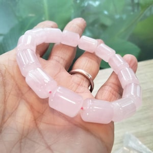 May include: A stretch bracelet composed of smooth, cylindrical, pale pink rose quartz beads. The beads are arranged in a continuous loop, connected by a thin, visible pink elastic cord that runs through the center of each polished stone bead.