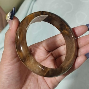May include: A brown bangle bracelet with a polished finish and a marbled effect. The bracelet has a smooth, rounded form and a warm, earthy colour. The bracelet is made of a translucent material.