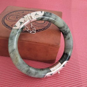 Könnte beinhalten: Ein Jade-Armreif mit silbernen Akzenten. Das Armband hat ein gesprenkeltes grün-graues Muster. Die silbernen Verschlüsse sind mit floralen Designs verziert und durch eine kleine Kette verbunden. Das Armband wird auf einer braunen Lederoberfläche präsentiert.