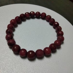 May include: A circular stretch bracelet composed of numerous faceted, deep maroon-colored beads. The beads are uniform in size and shape, featuring a polished finish that reflects light across their multiple angled surfaces in a consistent pattern.