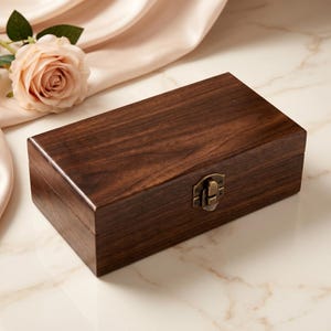 May include: A rectangular dark brown wooden box with a smooth, polished finish and a visible wood grain pattern. The box features a hinged lid secured by a small, antique-style brass-colored metal latch centered on the front panel.