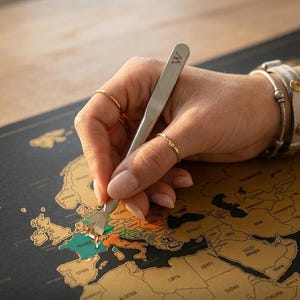 Op de afbeelding: Een persoon krabt een goudkleurige kras-af-kaart open met een zilveren gereedschap. De kaart toont Europa, met het woord "FRANCE" zichtbaar. Het gereedschap heeft de letter "W" gegraveerd. De kaart ligt op een houten oppervlak.