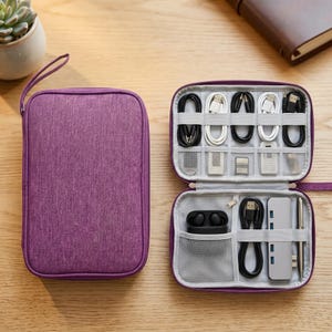Peut inclure: Un organisateur de voyage violet avec une poignée, ouvert pour révéler des compartiments avec des câbles, des hubs USB et d'autres accessoires technologiques. L'organisateur est sur une surface en bois.