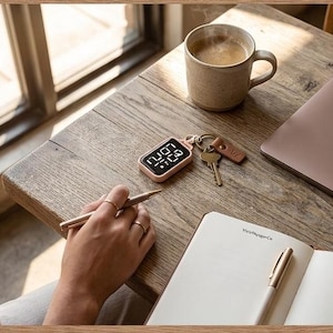 May include: A digital keychain with a rose gold case displays the time. A cup of coffee, keys, a laptop, a pen, and a notebook are on a wooden table. The notebook has the text "MakeHappyCo".