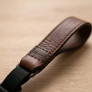 Peut inclure: Gros plan sur une sangle d'appareil photo en cuir marron avec une surface texturée. La sangle a une conception en boucle et est attachée à une sangle en nylon noir avec un anneau métallique. Le cuir est d'un brun foncé riche.