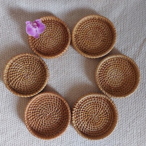 May include: Six round woven wicker coasters in a natural brown color arranged in a circle on a white textured surface. A single purple orchid is placed on top of one of the coasters.