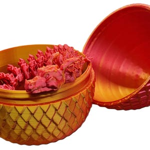 May include: A red and gold dragon egg-shaped container with a removable top. The top is open and reveals a pink dragon inside. The container is decorated with a scaly pattern.