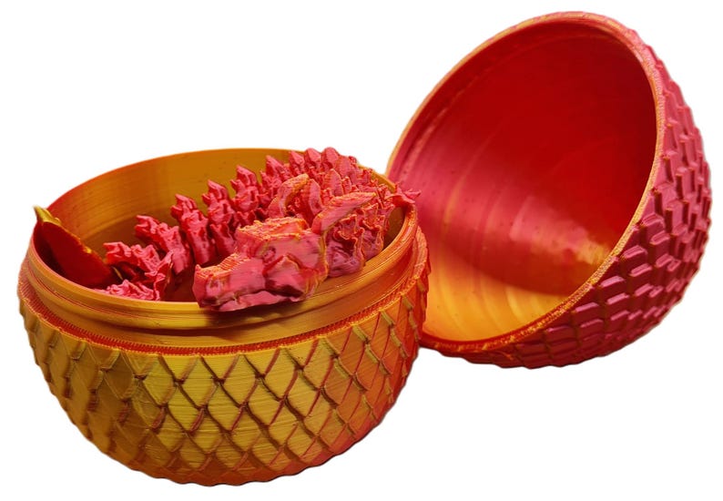 May include: A red and gold dragon egg-shaped container with a removable top. The top is open and reveals a pink dragon inside. The container is decorated with a scaly pattern.