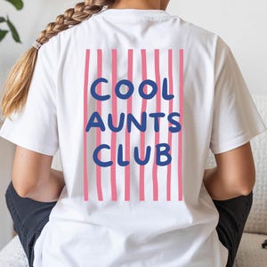May include: A white short-sleeved t-shirt featuring a graphic on the back. The design consists of vertical pink stripes with the text COOL AUNTS CLUB printed in bold blue letters over the stripes. The shirt has a crew neckline and short sleeves.