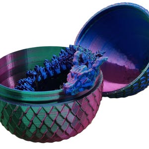 May include: A dragon egg-shaped container with a blue dragon sculpture inside. The container is made of a green and pink material with a textured surface. The dragon sculpture is blue and has a textured surface.