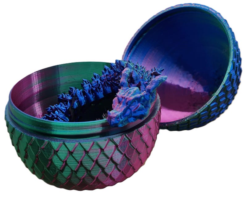 May include: A dragon egg-shaped container with a blue dragon sculpture inside. The container is made of a green and pink material with a textured surface. The dragon sculpture is blue and has a textured surface.