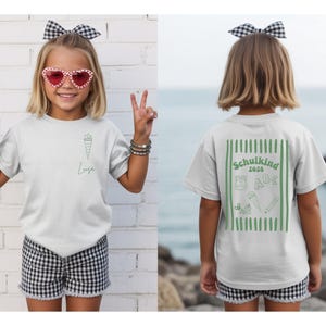 May include: A light gray short-sleeve t-shirt featuring a small green school cone graphic and the name Luisa on the front, with a large green graphic on the back displaying the text Schulkind 2026, a backpack, ABC letters, scissors, and a pencil.