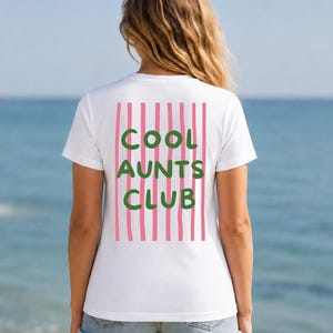 May include: A white short-sleeve t-shirt featuring a graphic on the back with vertical pink stripes and the text COOL AUNTS CLUB in green block letters. The shirt is paired with light blue denim shorts, showcasing a casual and simple design.