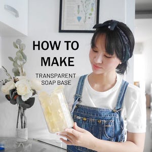 May include: A woman holding a clear glass beaker filled with transparent soap base cubes. The text "HOW TO MAKE TRANSPARENT SOAP BASE" is displayed in black capital letters. The woman is wearing denim overalls and a white t-shirt. A bouquet of white roses and greenery is in a vase.