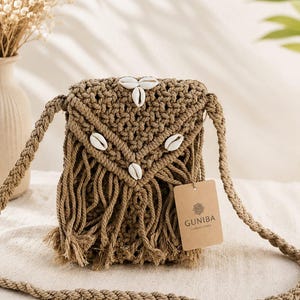 May include: A tan macrame crossbody bag featuring a triangular flap adorned with four white cowrie shells. The bag has a braided shoulder strap and a fringe bottom. A rectangular brown paper tag is attached, printed with the text GUNIBA HANDLOOMS.