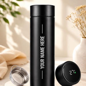 May include: A matte black cylindrical vacuum insulated water bottle featuring the white text YOUR NAME HERE vertically centered. The bottle includes a detachable metal tea infuser strainer and a digital LED temperature display on the lid.