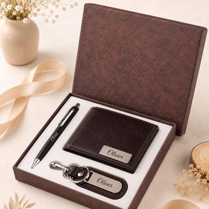 Può includere: Una scatola regalo marrone scuro contenente un set di accessori abbinati. Il set include una penna a sfera nera, un portafoglio pieghevole marrone scuro e un portachiavi marrone scuro. Ogni articolo presenta una targhetta in metallo argentato con inciso Oliver.