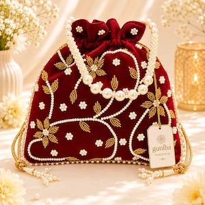 May include: A deep red velvet drawstring pouch bag featuring intricate gold thread embroidery and white pearl bead embellishments. The bag has a pearl bead handle and a hanging tag with the text guniba HANDLOOM. Gold tassels hang from the sides.