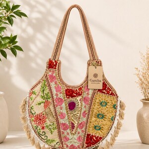 Puede incluir: Un bolso de mano en forma de media luna con un diseño de parches de bordados florales en rosa, rojo y dorado. El bolso tiene dos asas, un fleco de borlas beige en el borde inferior y una etiqueta con el nombre Guniba Handloom.