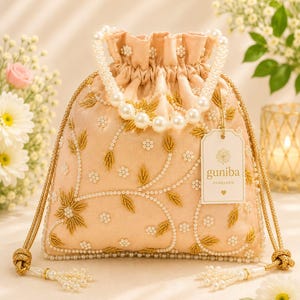 Puede incluir: Un bolso tipo saco de color melocotón con cordón, bordado con hilo dorado en motivos florales y detalles de perlas blancas. El bolso tiene un asa de perlas y cordones dorados con borlas de perlas. Una etiqueta dice guniba HANDLOOM.