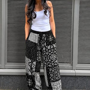 Könnte beinhalten: Eine beige Baseballkappe, ein weißes Tanktop und eine locker sitzende, schwarz-weiß gemusterte Hose mit einem Patchwork-Design aus verschiedenen geometrischen und floralen Motiven. Die Hose hat einen elastischen Bund mit Kordelzug und weiße Sneaker.